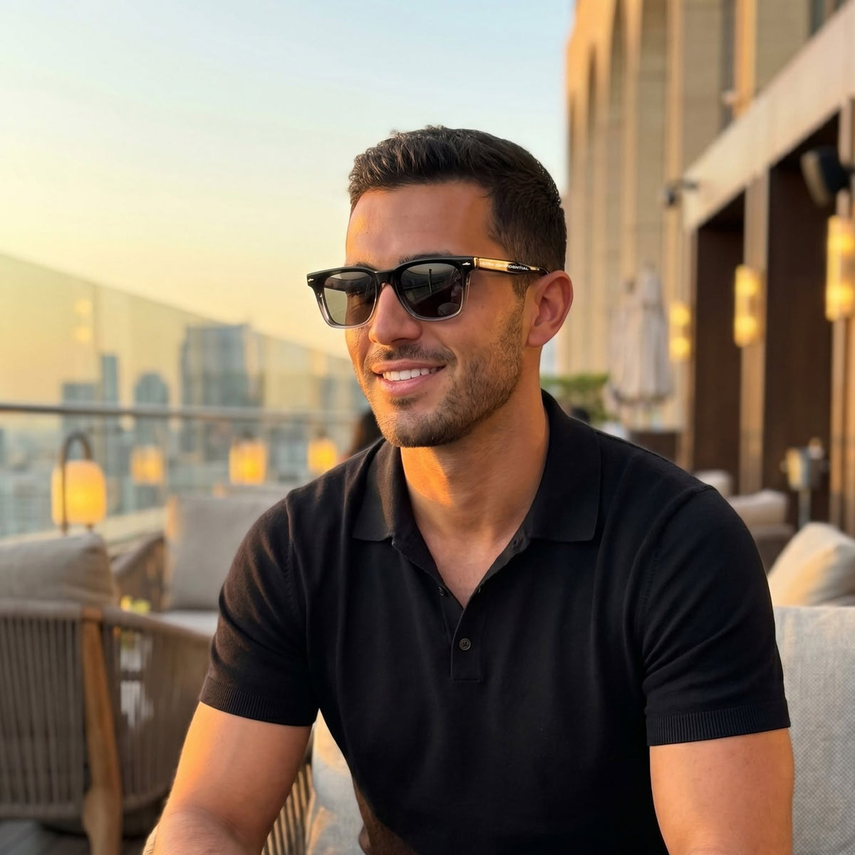 A man wearing black sunglasses with gold accents, a black polo shirt, and a gold watch sits on a rooftop patio holding a drink.